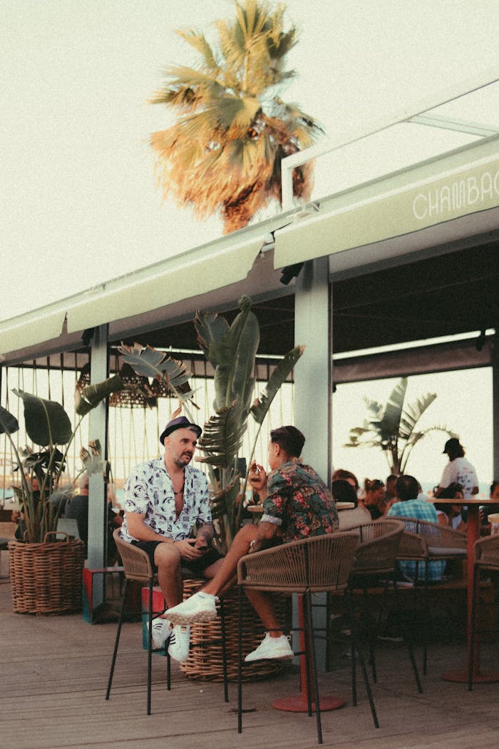 Two men enjoy a lively conversation at a tropical outdoor café, surrounded by lush greenery and warm summer vibes.
