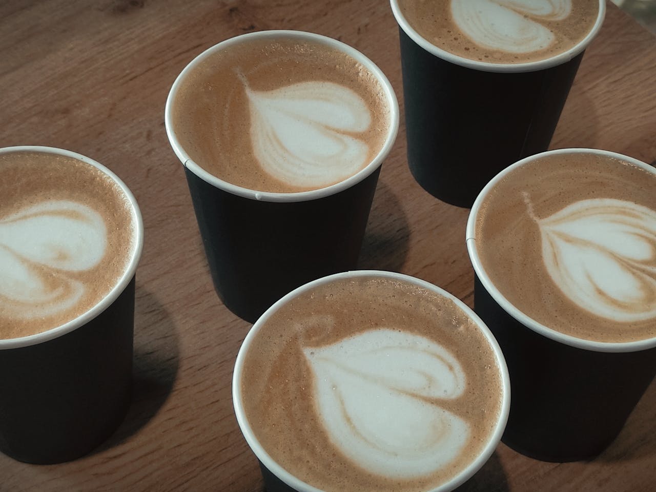 Top view of five lattes with heart latte art in black cups on wooden table.